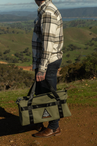 Person holding a green duffel bag with a mountainous landscape in the background