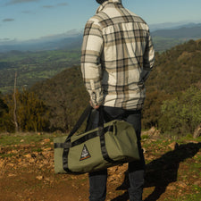 Person standing on a hill with a scenic view, holding a green duffel bag.