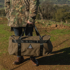 Person holding a brown duffel bag with a logo in a natural setting