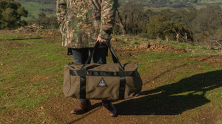 Person holding a brown duffel bag with a logo in a natural setting