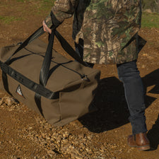 Person in camouflage holding a large brown bag outdoors on a dirt path