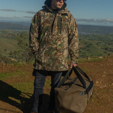 Person in camouflage jacket and beanie holding a duffel bag in a scenic outdoor setting