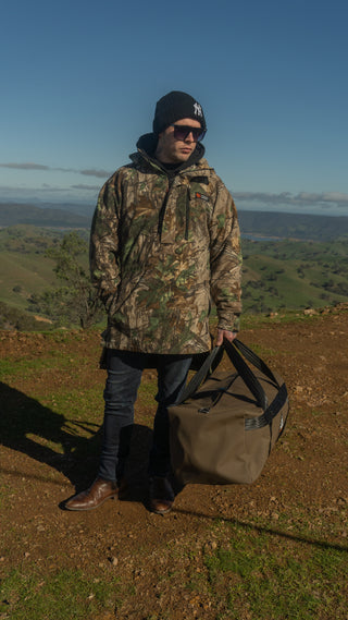 Person in camouflage jacket and beanie holding a duffel bag in a scenic outdoor setting