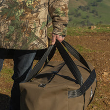 Person in camouflage jacket holding a large duffel bag with a scenic background