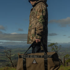Person in camouflage jacket holding a duffel bag with a logo, standing on a trail with a scenic background.
