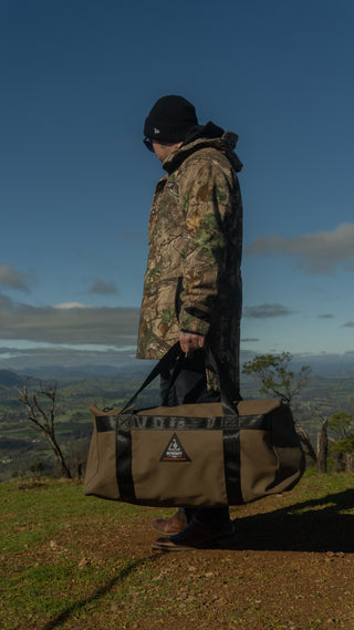 Person in camouflage jacket holding a duffel bag with a logo, standing on a trail with a scenic background.