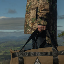 Person holding a camouflage jacket and a large duffel bag with a mountainous landscape in the background.
