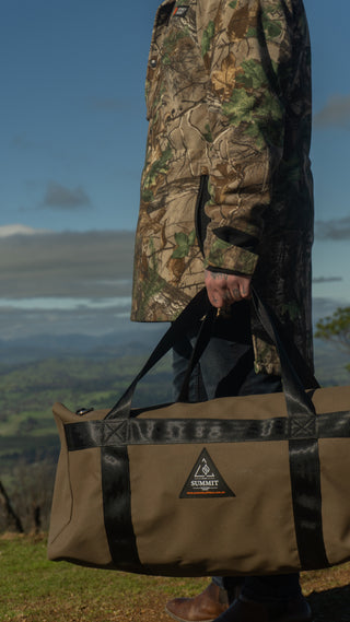 Person holding a camouflage jacket and a large duffel bag with a mountainous landscape in the background.