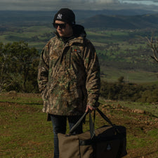 Person in camouflage jacket and sunglasses holding a bag in a scenic outdoor setting with mountains and fields.