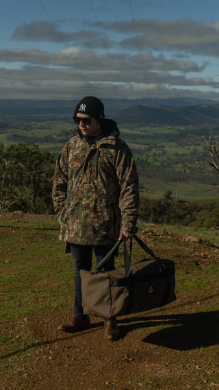 Person in camouflage jacket and sunglasses holding a bag in a scenic outdoor setting with mountains and fields.