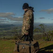 Person in camouflage jacket holding a bag with a scenic landscape in the background