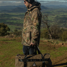 Person in camouflage jacket holding a duffel bag in a scenic outdoor setting with mountains and greenery.