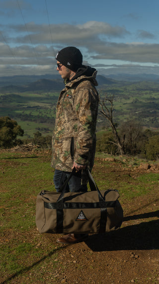 Person in camouflage jacket holding a duffel bag in a scenic outdoor setting with mountains and greenery.