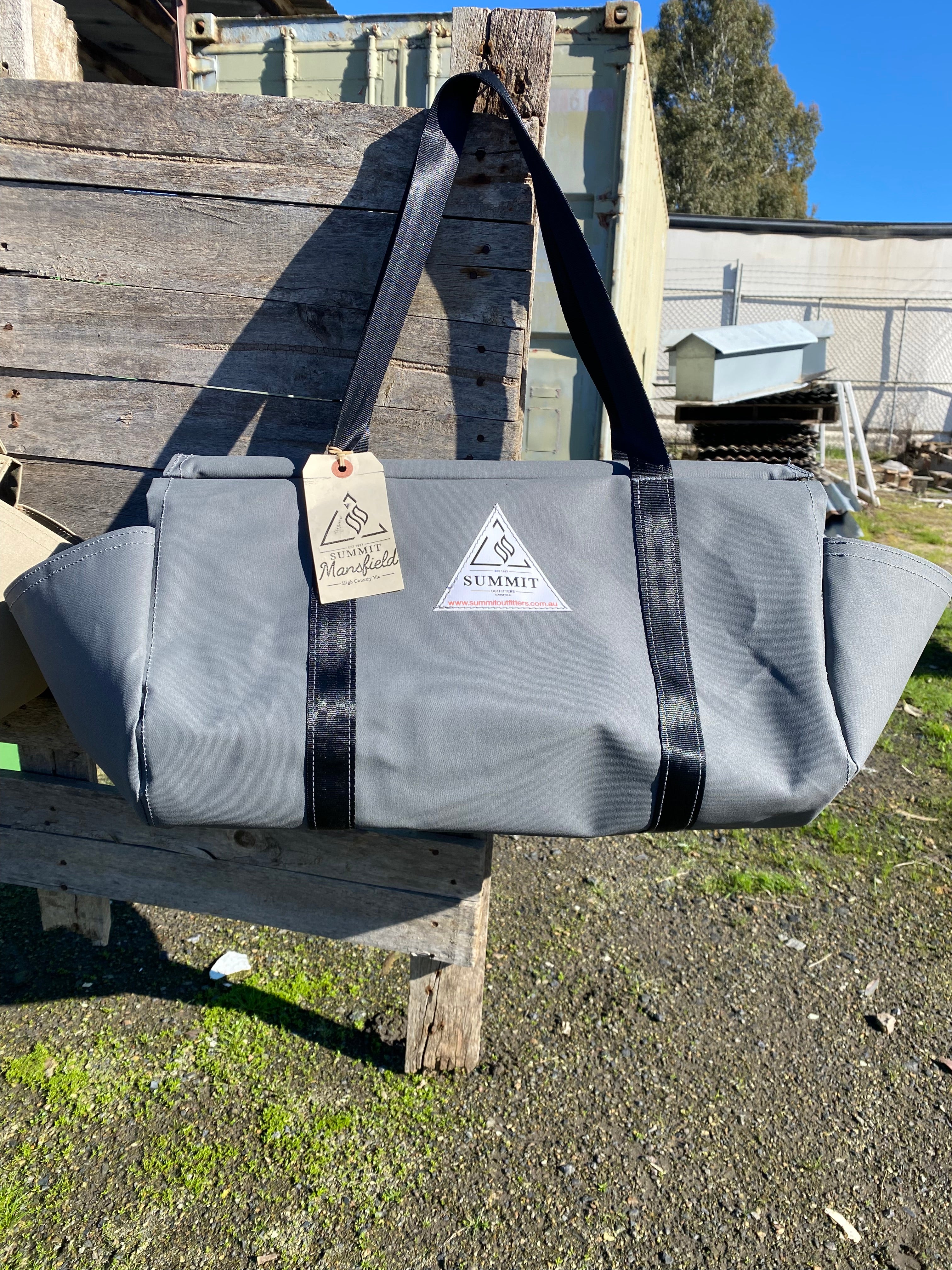 grey coloured canvas firewood carrier bag with black strapped handles and small triangle summit outfitters pvc logo patch on front of product, hanging off wooden palette