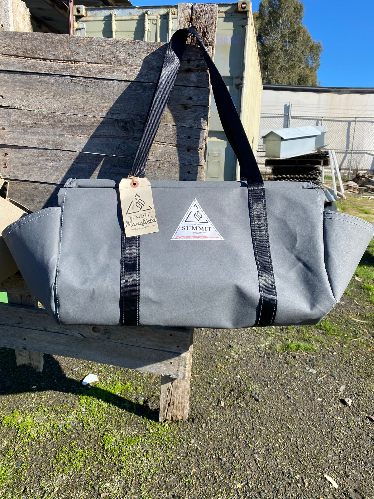 grey coloured canvas firewood carrier bag with black strapped handles and small triangle summit outfitters pvc logo patch on front of product, hanging off wooden palette
