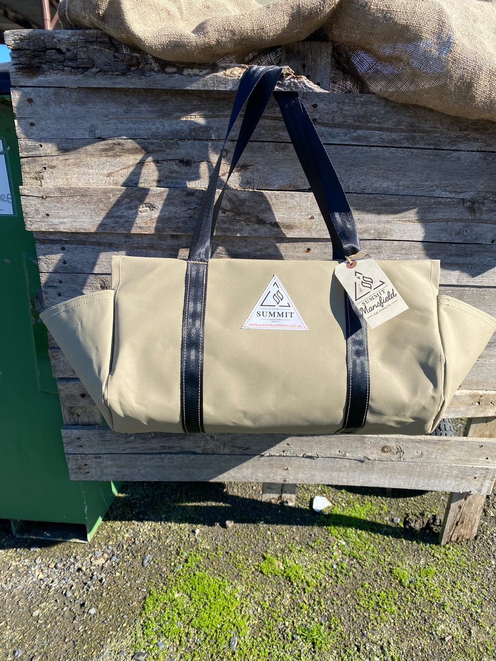 beige coloured canvas firewood carrier bag with black strapped handles and small triangle summit outfitters pvc logo patch on front of product, hanging off wooden palette