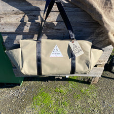 beige coloured canvas firewood carrier bag with black strapped handles and small triangle summit outfitters pvc logo patch on front of product, hanging off wooden palette