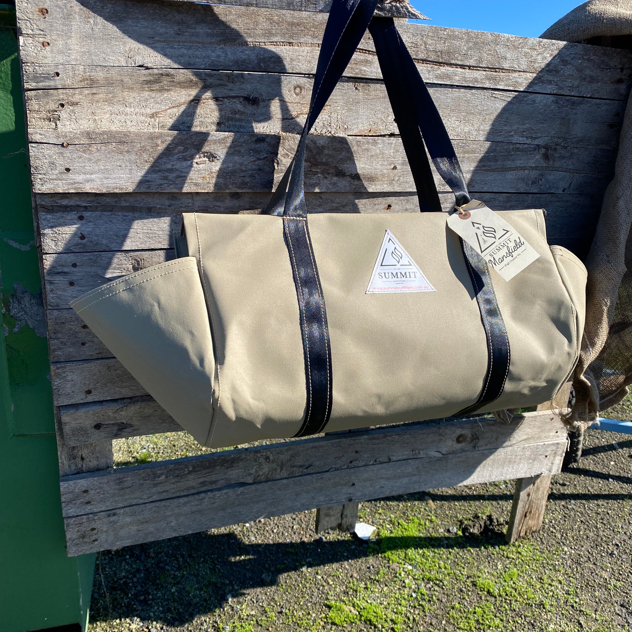 beige coloured canvas firewood carrier bag with black strapped handles and small triangle summit outfitters pvc logo patch on front of product, hanging off wooden palette