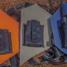 Three radio holsters in orange, sand, and blue with radio holders on a pine needle covered floor
