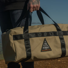 Person holding a beige duffel bag with a logo against a scenic background