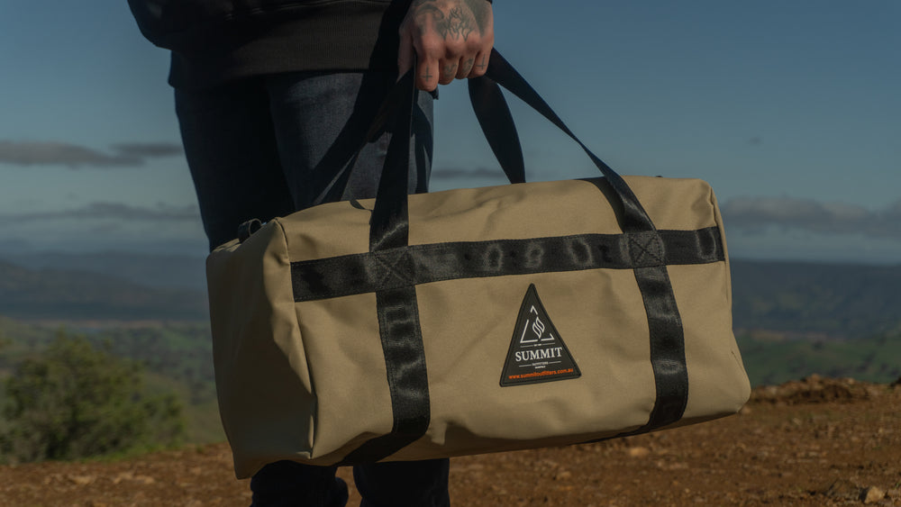 Person holding a beige duffel bag with a logo against a scenic background