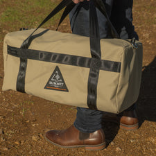 Person holding a beige duffel bag with black straps on a dirt ground