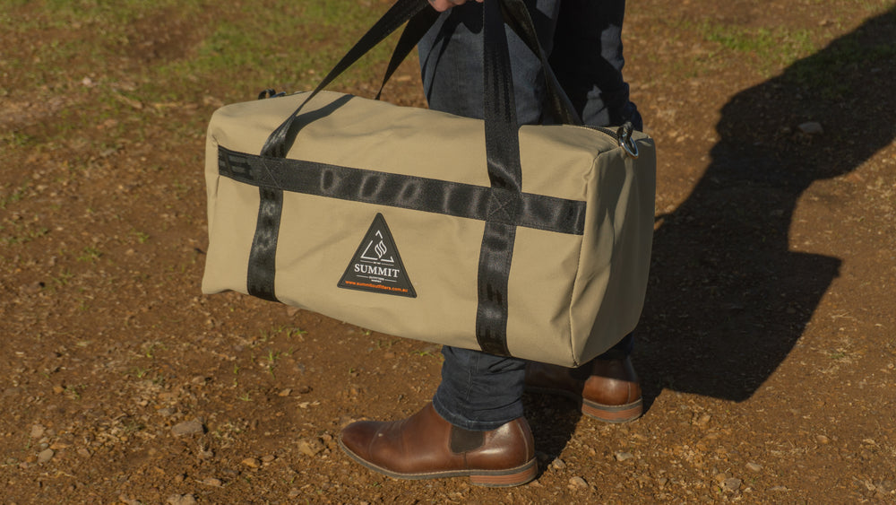 Person holding a beige duffel bag with black straps on a dirt ground