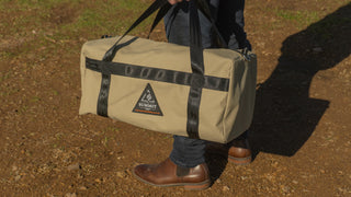 Person holding a beige duffel bag with black straps on a dirt ground