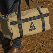 Person holding a beige duffel bag with black straps on a dirt ground