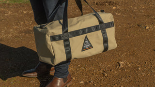 Person holding a beige duffel bag with black straps on a dirt ground