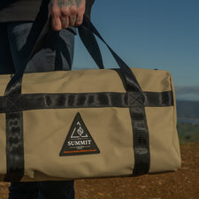 Person holding a beige duffel bag with black straps against a scenic background