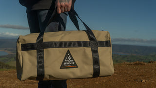 Person holding a beige duffel bag with black straps against a scenic background